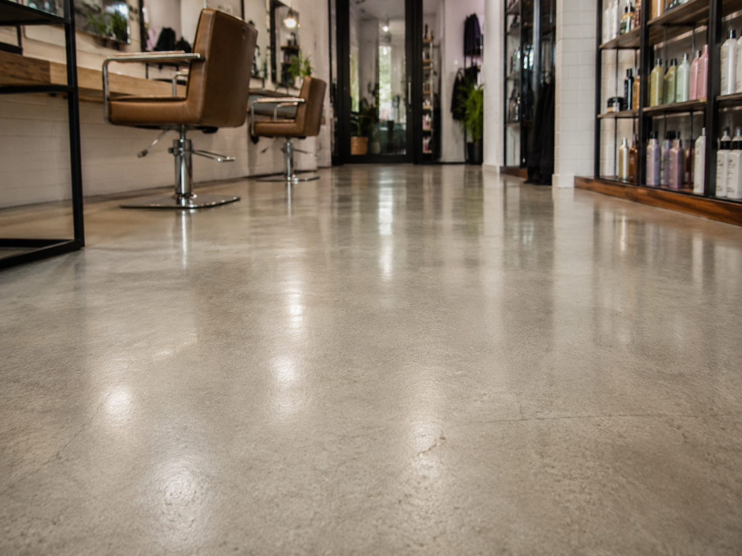 Close-up of a polished concrete floor coating inside a modern salon, highlighting a smooth, easy-to-clean surface with styling chairs and product shelves in the background.